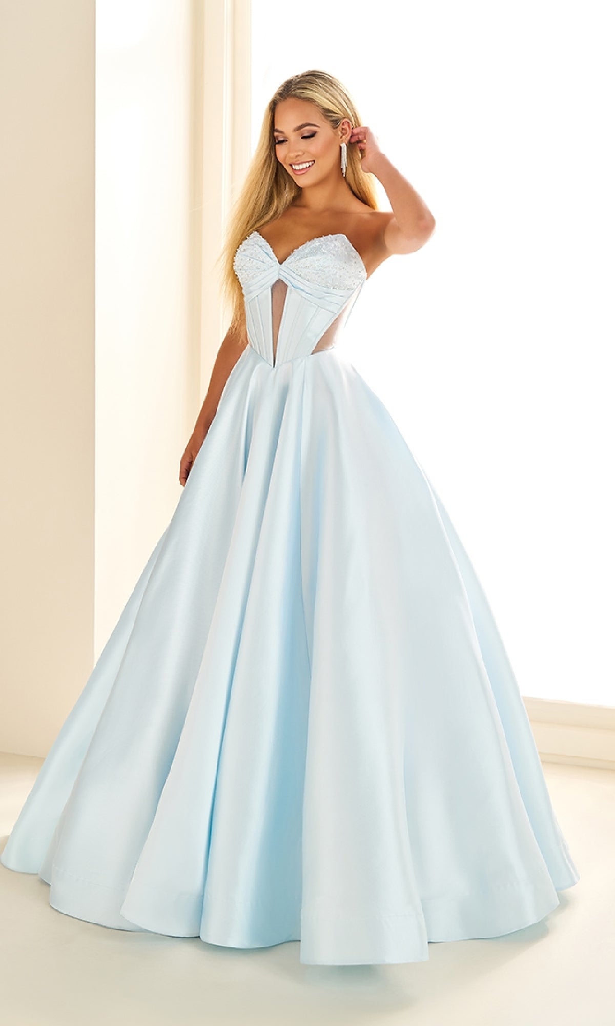 A woman wears the Ellie Wilde EW36025, a light blue satin long prom dress with a corset bodice and full skirt, standing indoors near a window.
